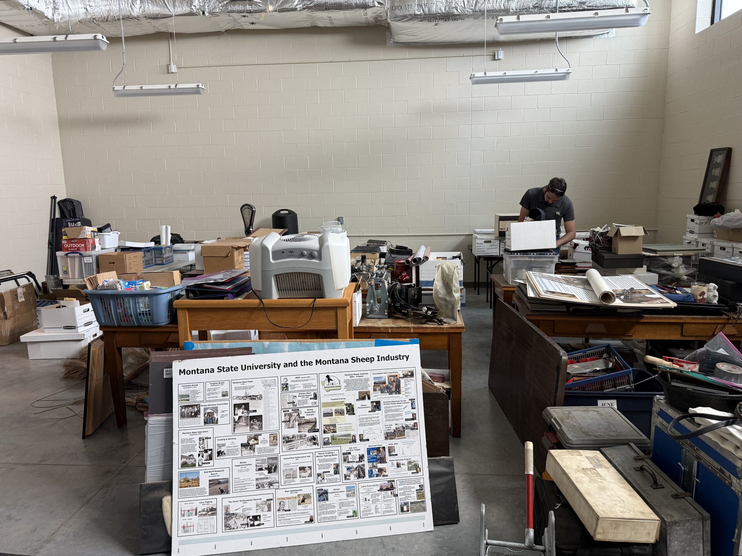 Messy workshop with a man at a cluttered workstation on the right and a large Montana Sheep Industry poster in the foreground.