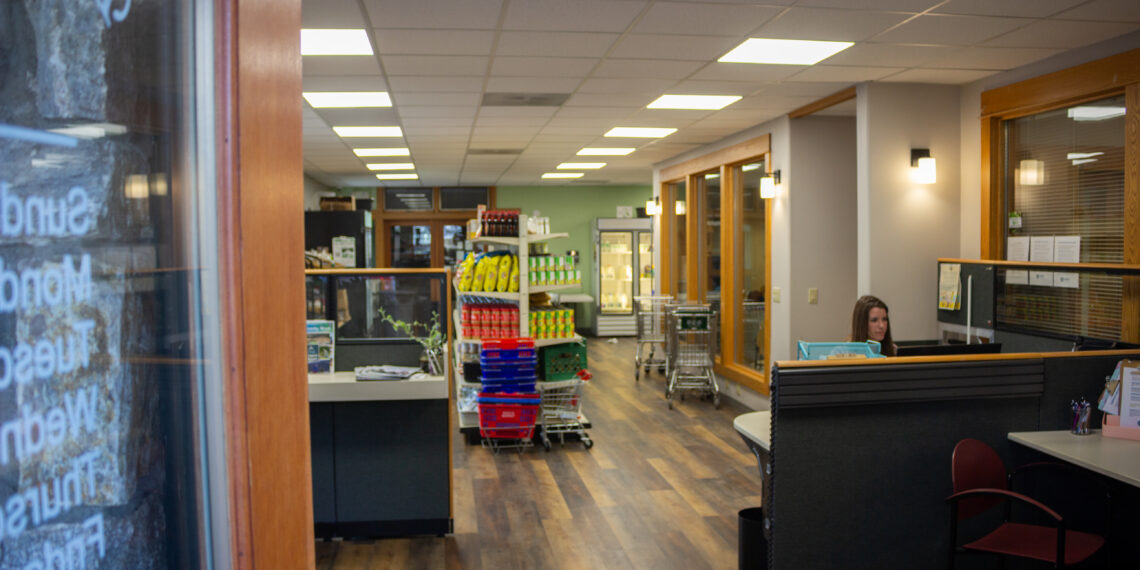 Interior of a store seen from the doorway: checkout desks on the right, central shelves with snacks, and a woman seated at a desk near the far right.