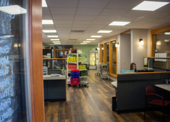 Interior of a store seen from the doorway: checkout desks on the right, central shelves with snacks, and a woman seated at a desk near the far right.