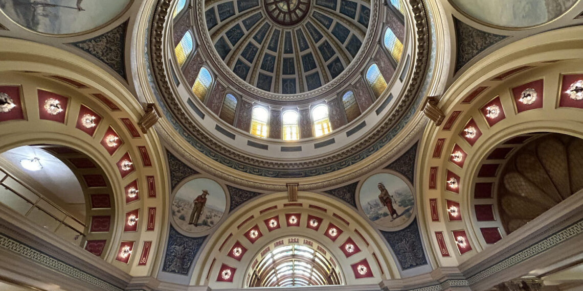 Ornate domed ceiling in a grand hall, featuring a blue segmented dome with skylights and decorative arches with statues.