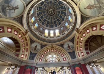 Ornate domed ceiling in a grand hall, featuring a blue segmented dome with skylights and decorative arches with statues.