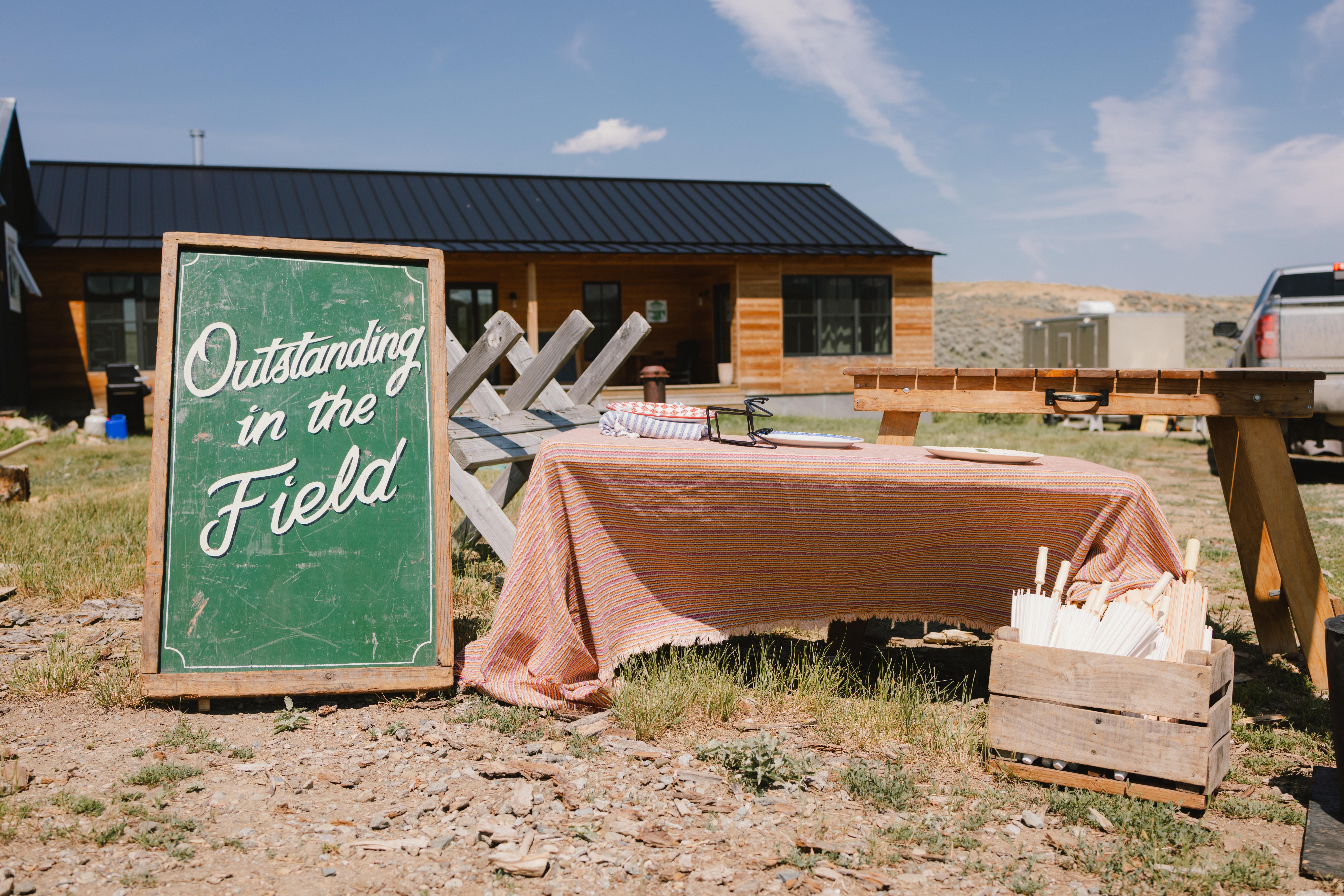 Outdoor dining setup with a striped tablecloth and utensils in front of a wooden building, beside a green chalkboard sign reading 'Outstanding in the Field'.
