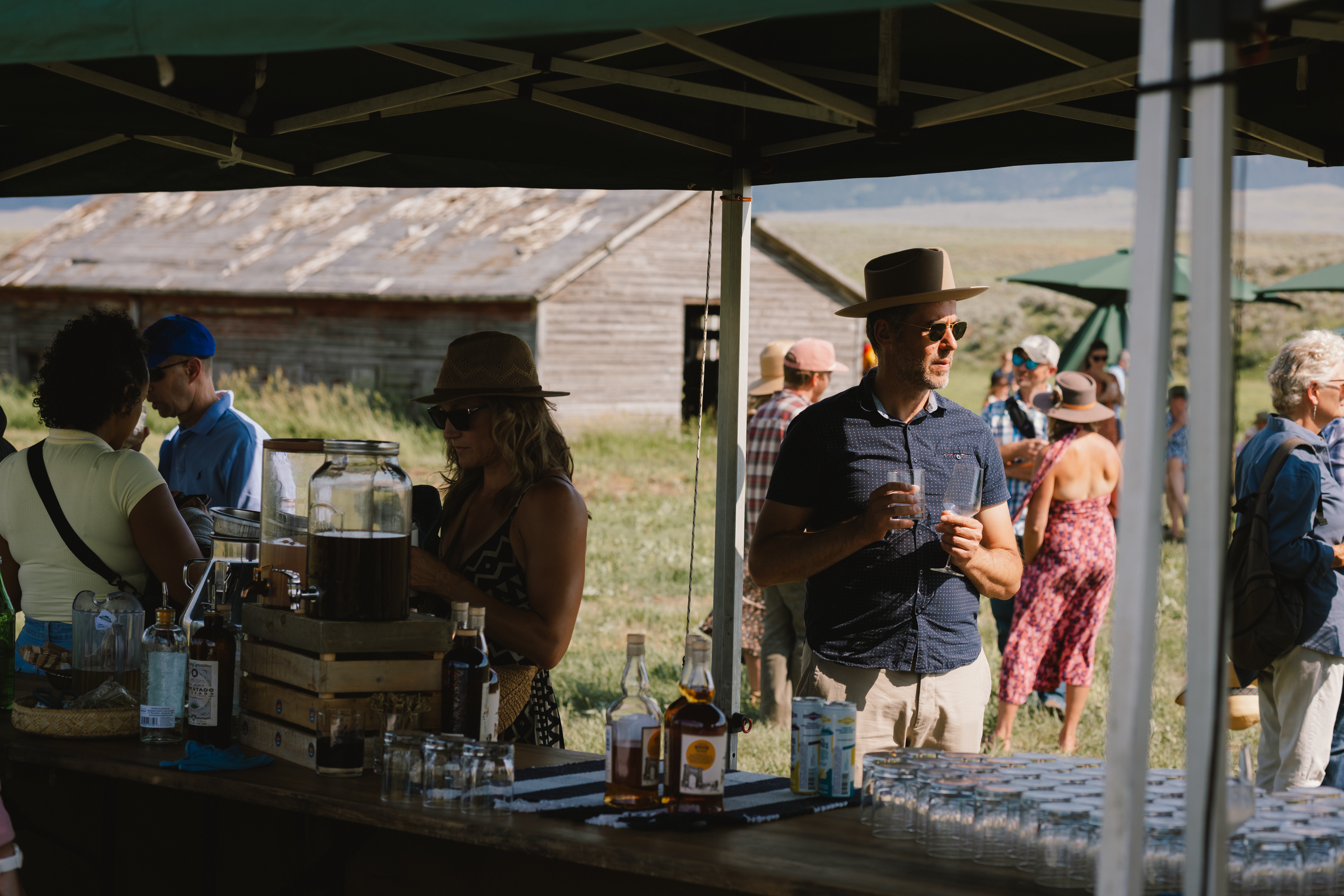 Outdoor sake: people mingle under a canopy at a rustic outdoor drinks stand with bottles and jars on the table