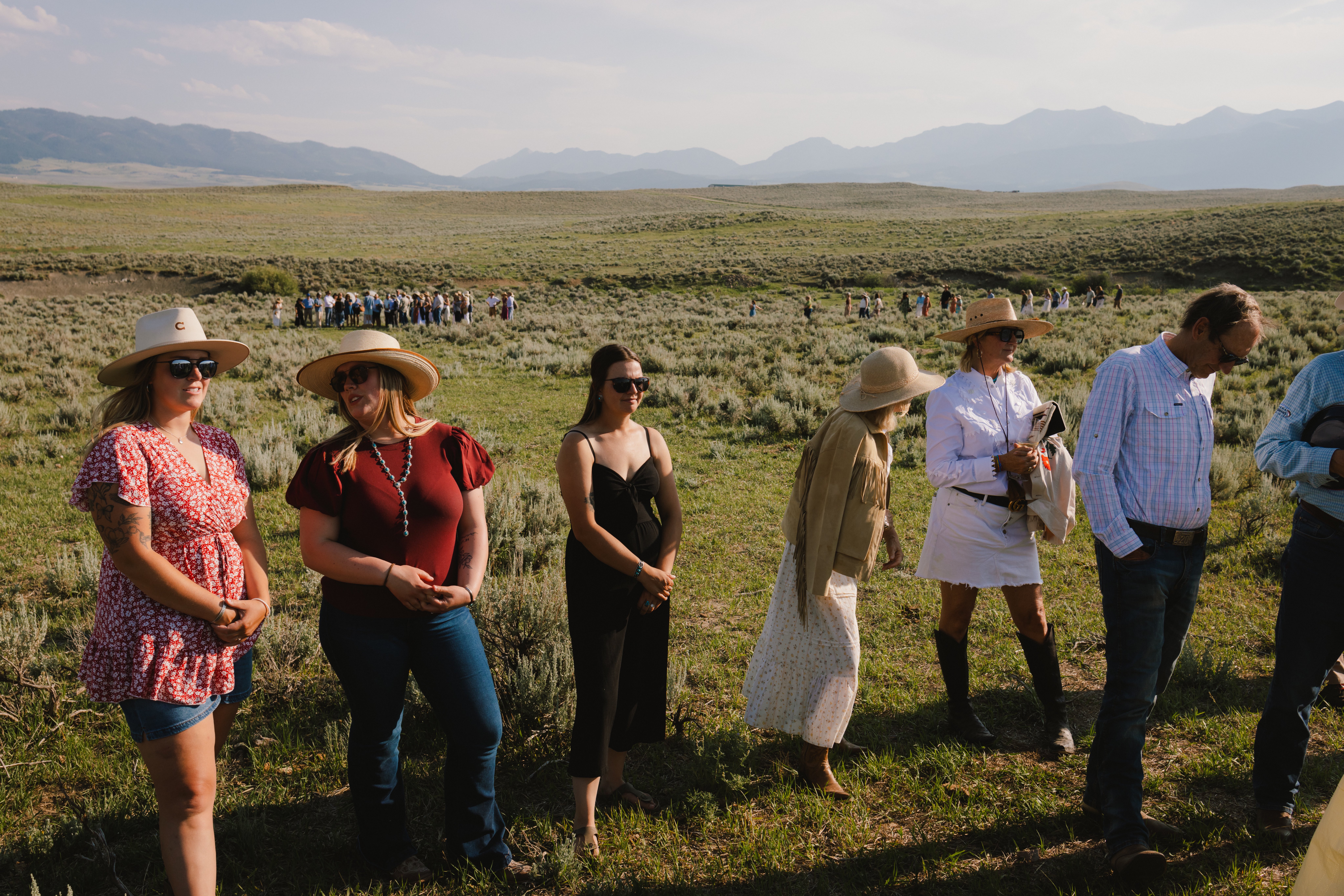 Group of people standing in a wide open sagebrush plain with mountains in the distance on a sunny day; foreground individuals wear hats and sunglasses, chatting casually.