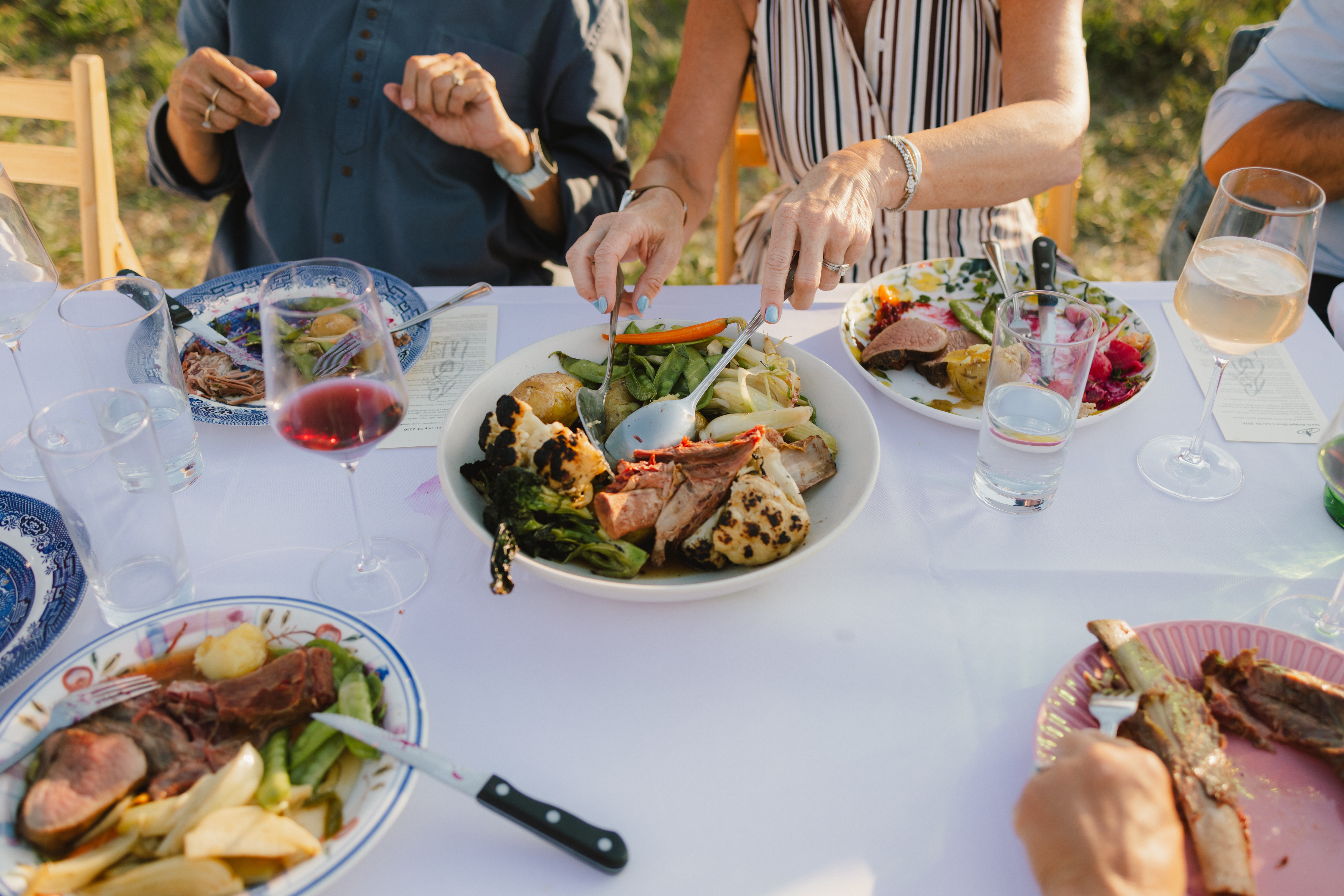 Outdoor family-style dinner with hands reaching to share a plate of roasted vegetables and meat, wine glasses on a white tablecloth.