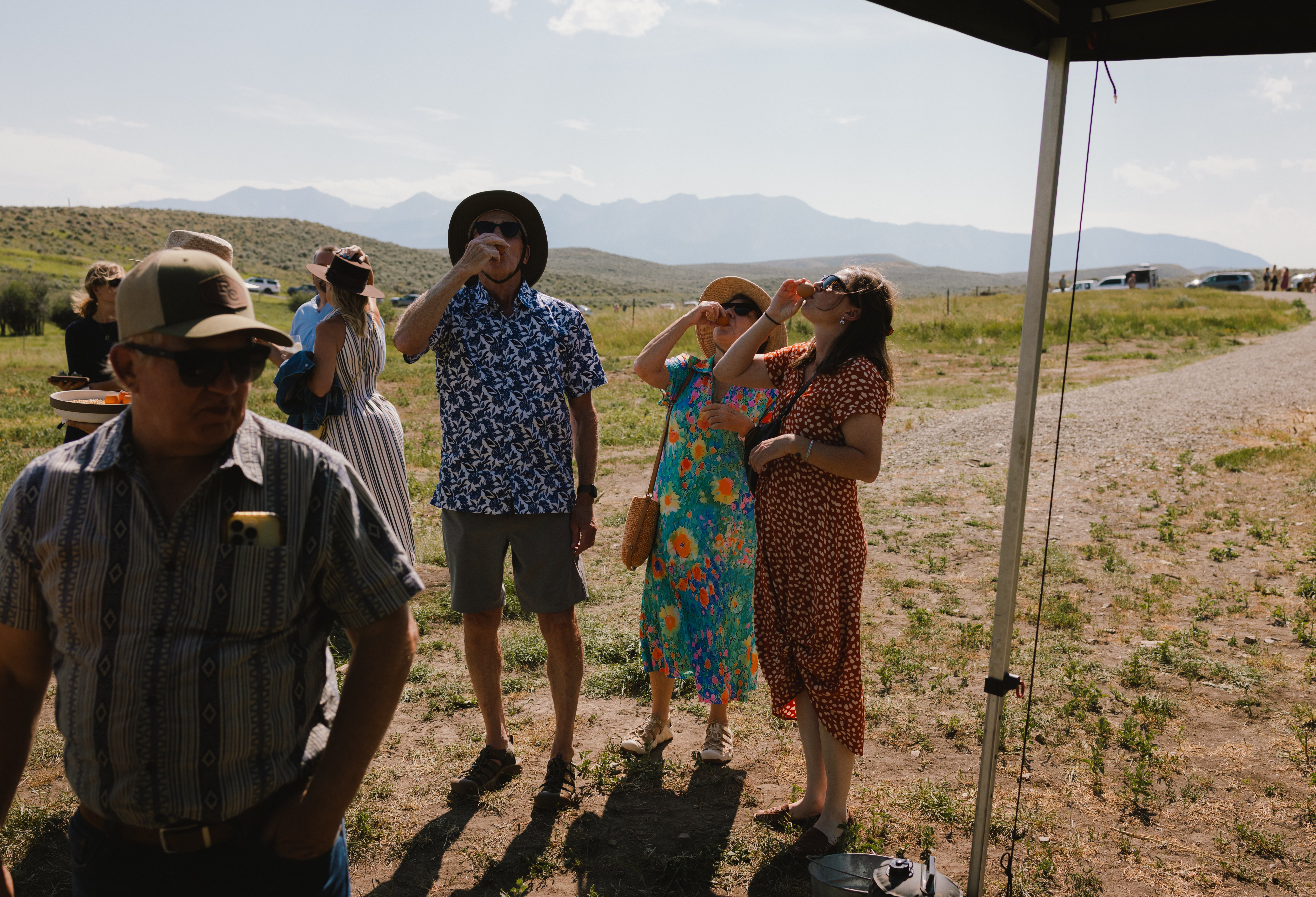 Group of people outdoors at a sunny rural gathering, some drinking from cups, wearing hats and colorful dresses, with mountains in the distance.