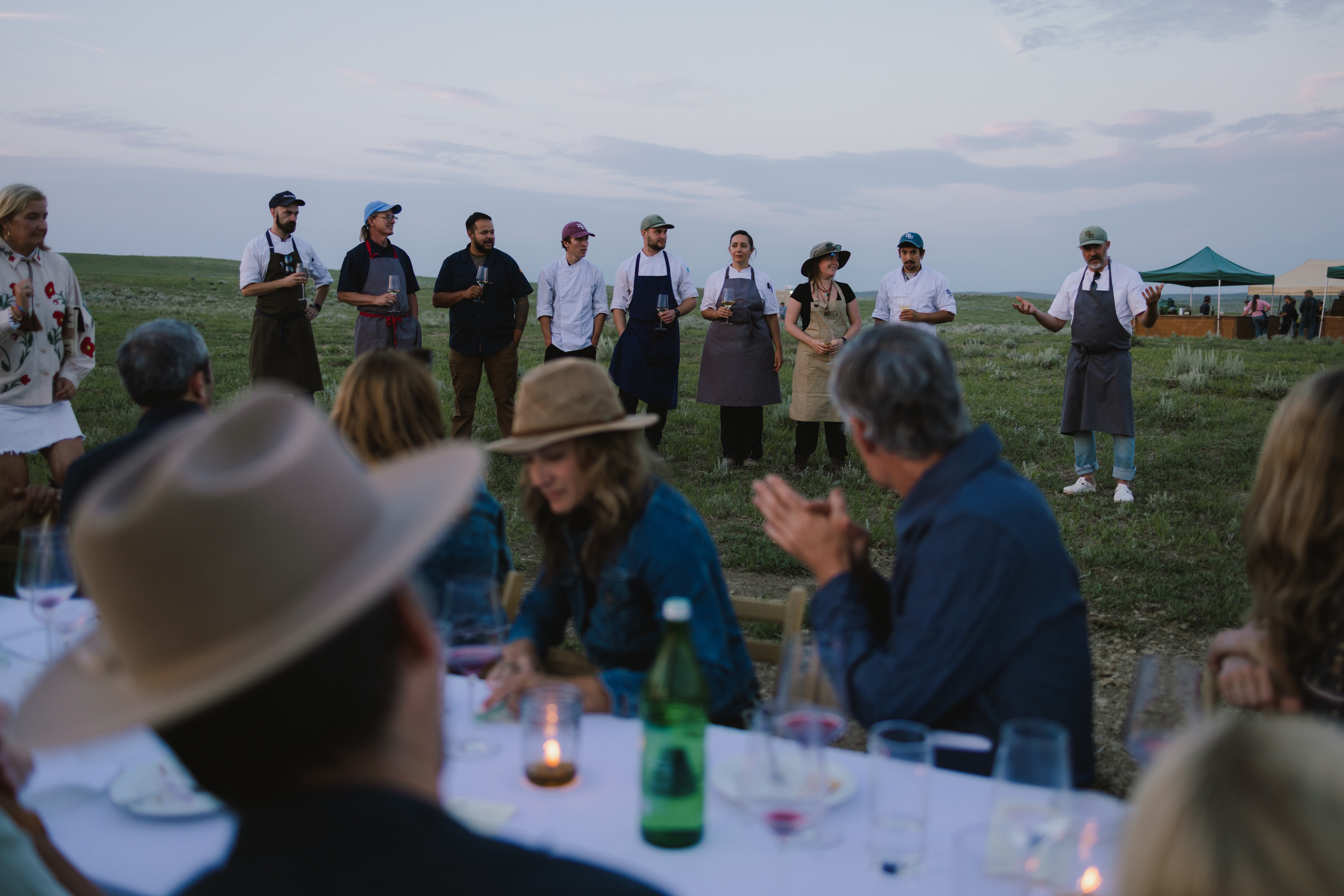 Line of people in aprons standing with wine glasses outdoors in a grassy field at dusk, tents in the background.