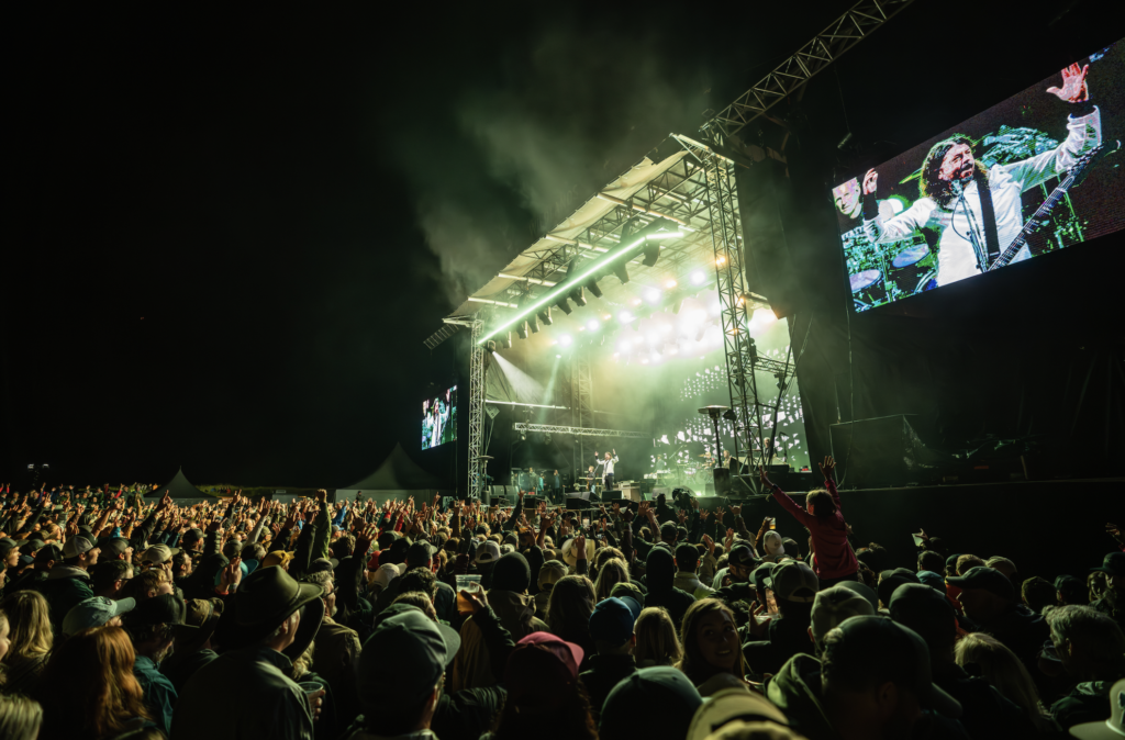 Large outdoor concert at night with a packed crowd raising hands toward a brightly lit stage and giant screen showing performers.