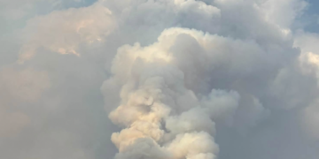 Massive plume of smoke rising from a wildfire over a mountain range, with a forested foreground and open plains.