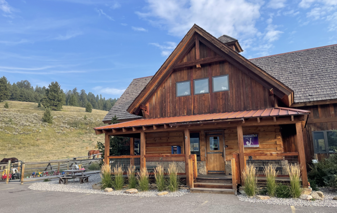 Rustic wooden lodge with a steep gabled roof and wraparound porch, set in a rural yard with a hillside and horses in the background.