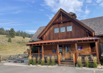 Rustic wooden lodge with a steep gabled roof and wraparound porch, set in a rural yard with a hillside and horses in the background.