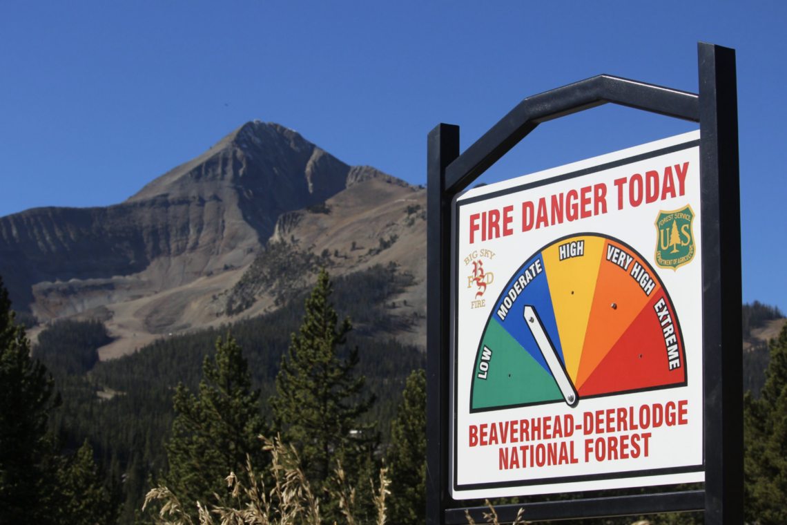 Sign reads 'FIRE DANGER TODAY' for Beaverhead-Deerlodge National Forest with a multi-color danger gauge and mountains in the background, under a blue sky.