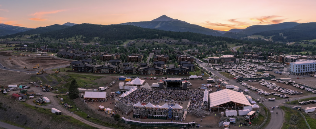Aerial view of a mountain town at sunset with a large outdoor concert, stage, tents, and a crowd gathered in a festival ground.