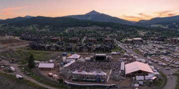 Aerial view of a mountain town at sunset with a large outdoor concert, stage, tents, and a crowd gathered in a festival ground.