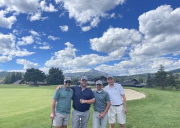 Four men in golf attire posing on a green golf course with mountains and houses in the background under a partly cloudy sky.