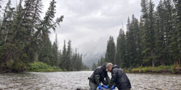 Two people in black waterproof jackets and gloves stand chest-deep in a river, handling equipment with a forested, cloudy backdrop.