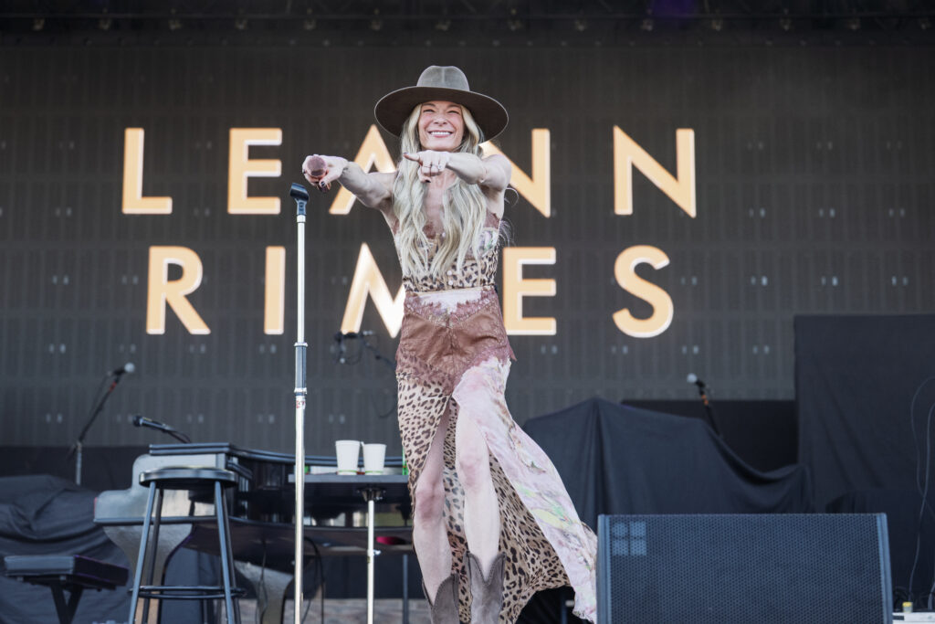 Female performer on stage wearing a wide-brim hat and leopard-print dress, holding a microphone and pointing to the audience against a backdrop of large letters behind her.