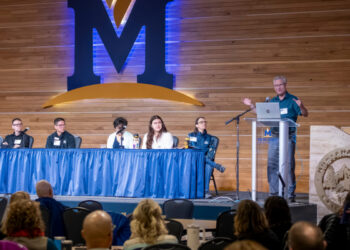 Panel discussion on a stage with five speakers seated behind a blue-draped table, a speaker at a podium with a laptop, and a large M logo on a wooden backdrop behind them.