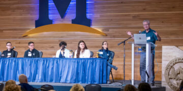 Panel discussion on a stage with five speakers seated behind a blue-draped table, a speaker at a podium with a laptop, and a large M logo on a wooden backdrop behind them.