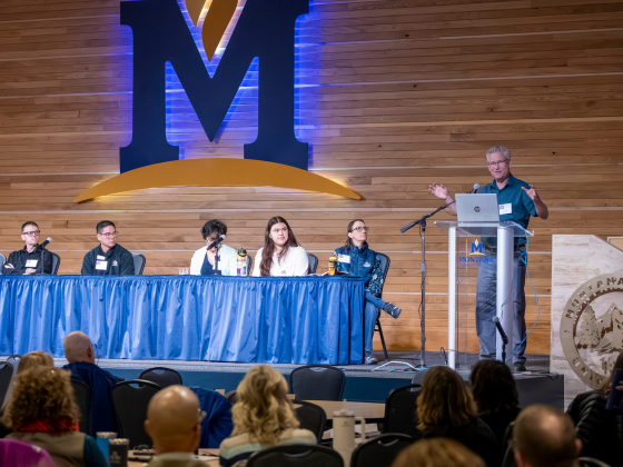 Panel discussion on a stage with five speakers seated behind a blue-draped table, a speaker at a podium with a laptop, and a large M logo on a wooden backdrop behind them.