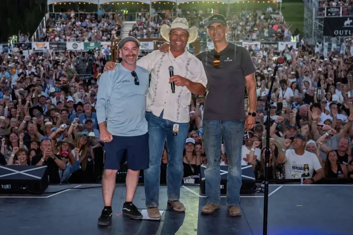 Three men stand on a stage posing for the crowd; center man in a white cowboy shirt and hat holds a microphone, flanked by two others in casual shirts while a large cheering audience watches.