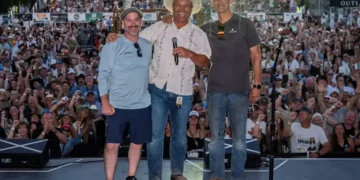 Three men stand on a stage posing for the crowd; center man in a white cowboy shirt and hat holds a microphone, flanked by two others in casual shirts while a large cheering audience watches.