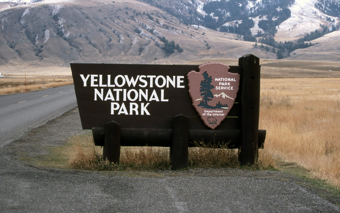 Yellowstone National Park sign with National Park Service emblem beside a rural road and mountainous backdrop.
