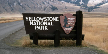 Yellowstone National Park sign with National Park Service emblem beside a rural road and mountainous backdrop.